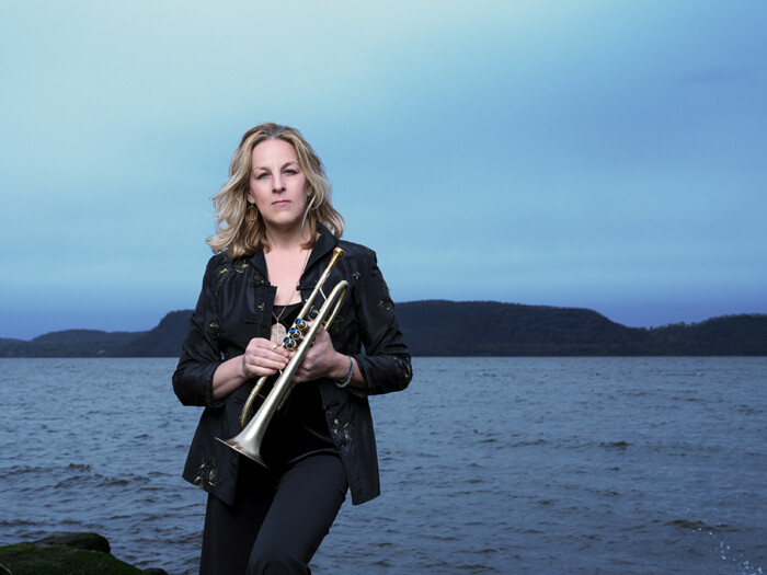 Porträtt av trumpetaren Ingrid Jensen, hon står vid vattnet i skymningen med trumpeten i sin hand